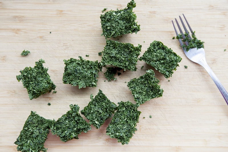 Nine cubes of chopped greens, possibly herbs or kale, are arranged on a wooden surface with a metal fork to the right. Small green bits are scattered around the cubes.