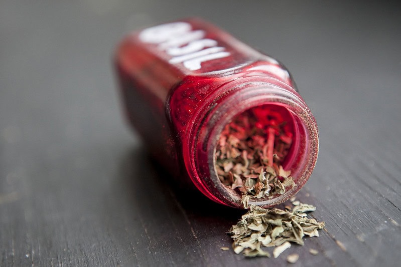A red spice jar labeled Basil lies on its side, spilling dried basil leaves onto a dark surface.