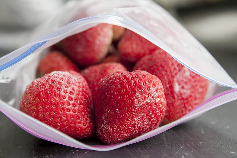 A close-up of several frozen strawberries inside a partially open clear plastic zipper bag on a dark surface.