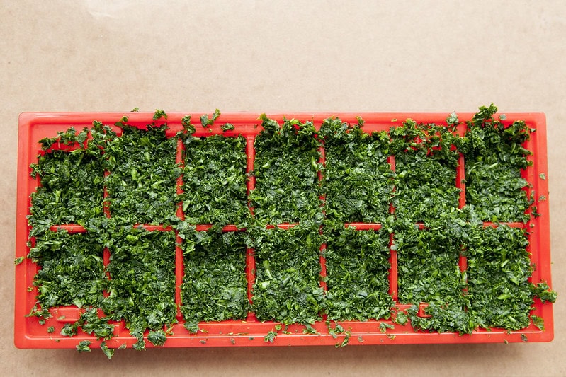 A red ice cube tray is filled with finely chopped fresh herbs, likely parsley or cilantro, on a beige surface.