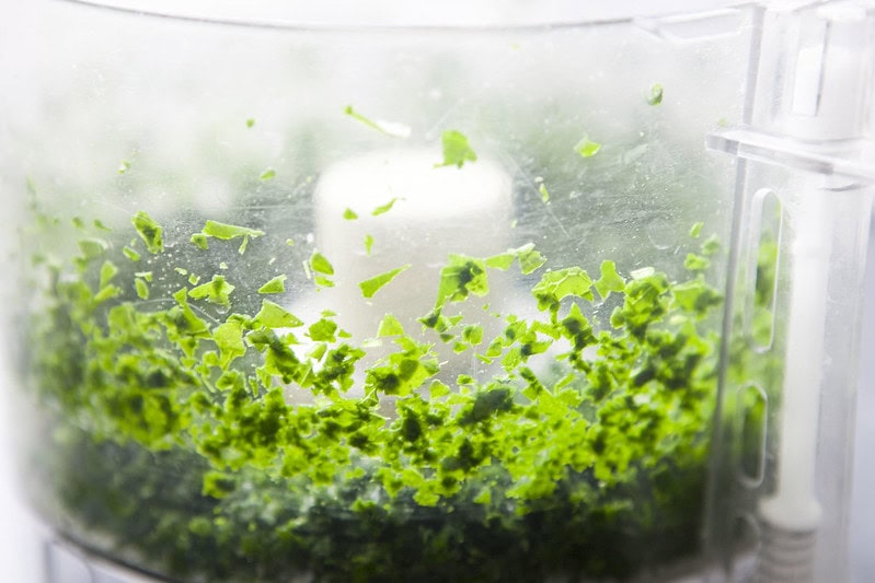 Chopped green herbs inside a food processor, with small bits sticking to the sides of the transparent container.