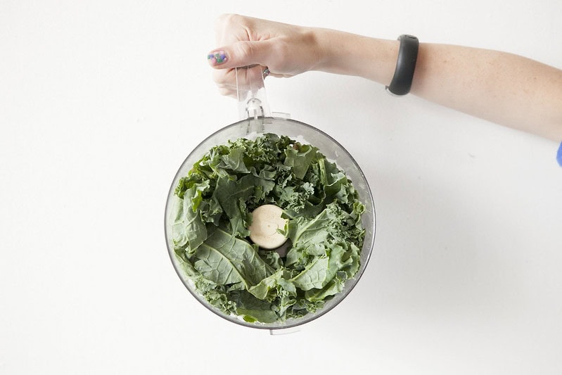 A hand holds a food processor bowl filled with fresh kale leaves, ready for processing, against a plain white background.