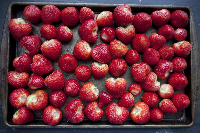 A baking tray filled with fresh, ripe strawberries, some with their tops removed, spread out in a single layer on a dark surface.