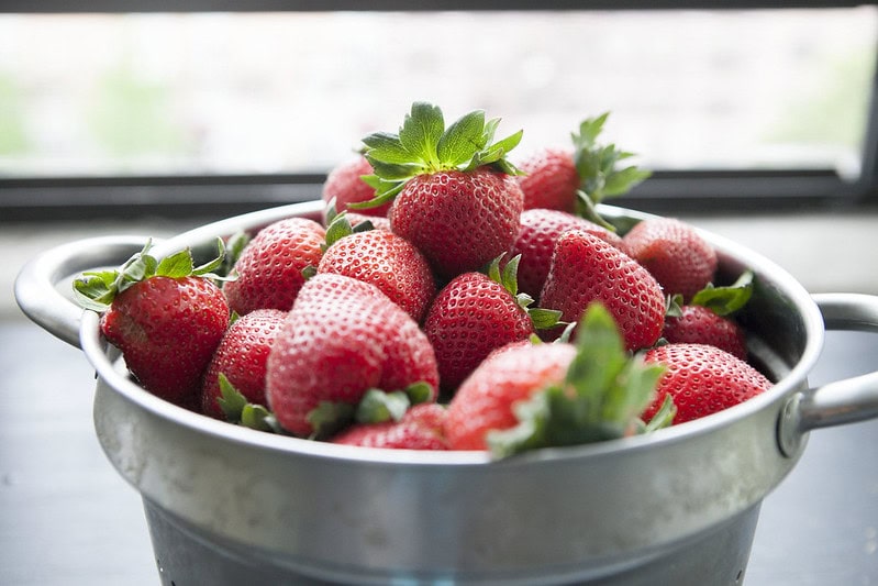 A metal colander filled with fresh, ripe strawberries sits on a dark surface, with a blurred window background letting in natural light.
