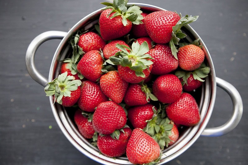 A metal colander filled with fresh, bright red strawberries sits on a dark, textured surface.