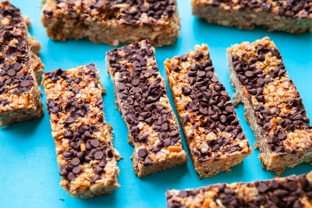 Eight granola bars with chocolate chips on top are arranged on a bright blue surface. The bars have a golden oat base and visible layers of chocolate chips spread across the tops.