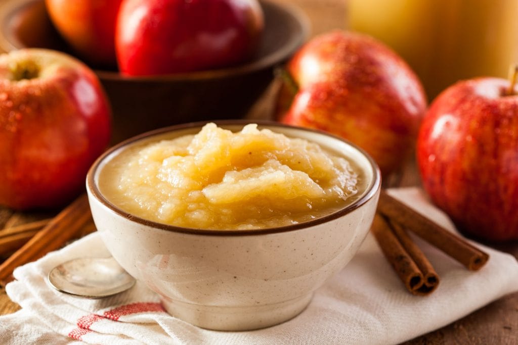 white bowl of applesauce with red apples in background next to cinnamon sticks