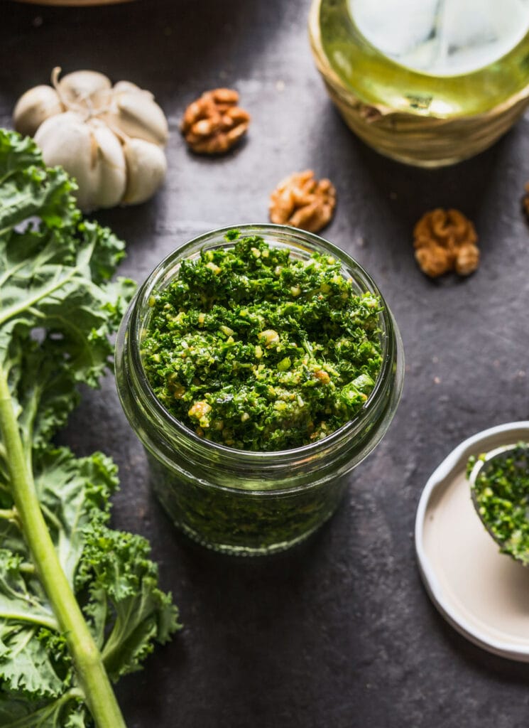 glass jar filled with kale pesto with other ingredients behind it on table. 