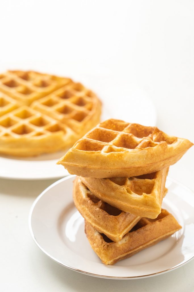 stack of 4 waffle pieces on plate with waffle in background.