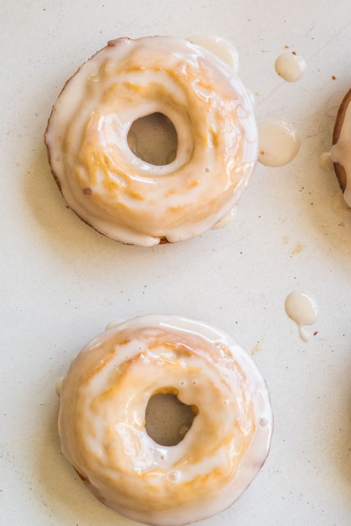 two donuts sitting on white table, glaze drippings on table too. 