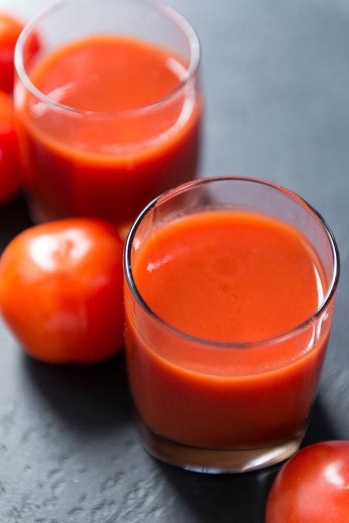 smooth tomato juice in glass with tomatoes behind it.