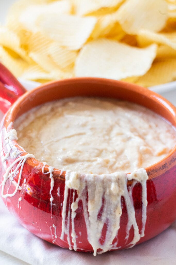 bowl of creamy onion dip with potato chips in the background.