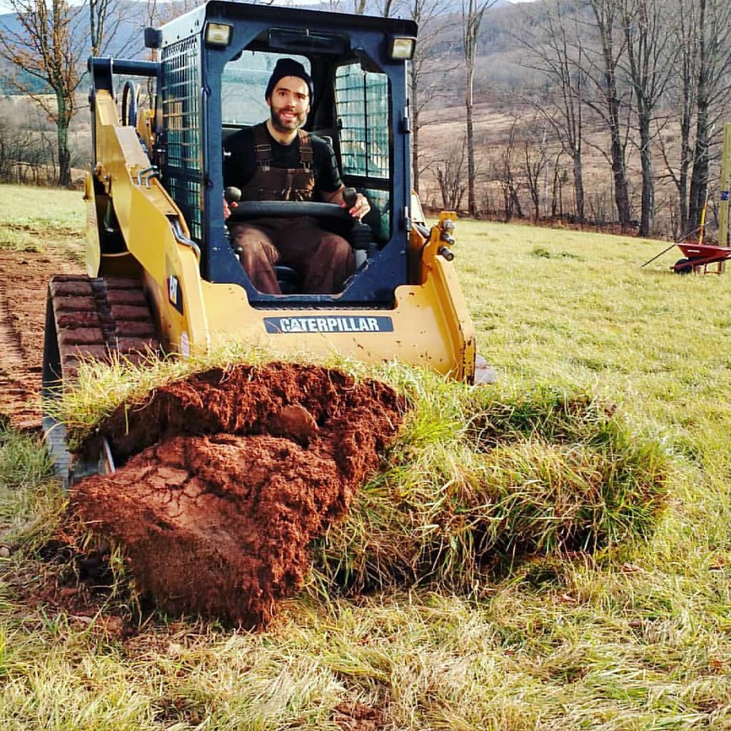 A man operates a Caterpillar skid steer loader, lifting a large chunk of grass and soil in a grassy field with bare trees and hills in the background.
