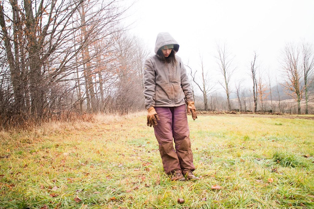 A person in a hooded jacket, gloves, and brown pants stands on a grassy field with bare trees in the background on a cloudy day.