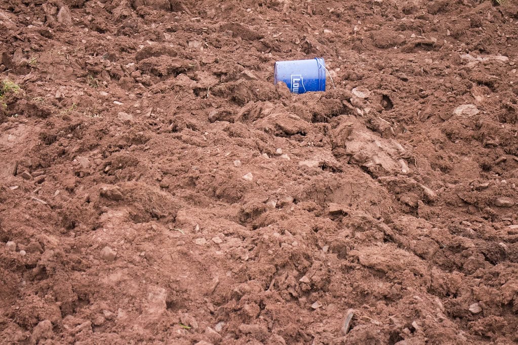A blue plastic container lies on uneven, brown dirt, surrounded by scattered rocks and clumps of soil.