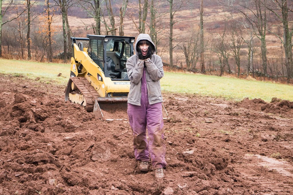 A person in a hooded jacket and gloves stands on muddy ground in front of a yellow bulldozer, with bare trees and a grassy hillside in the background.