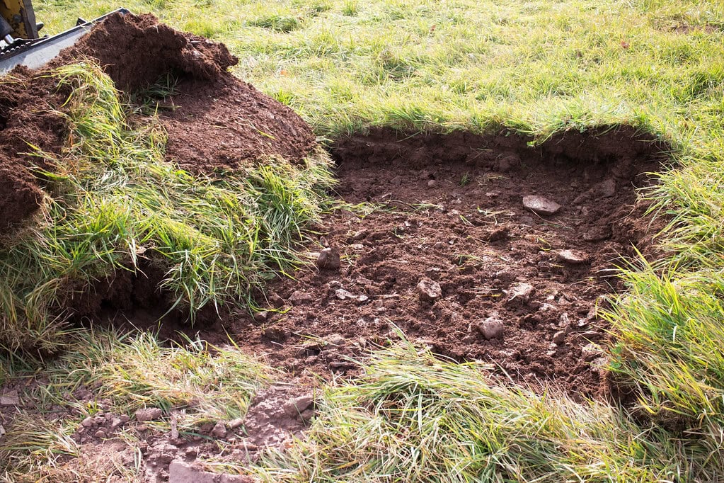 A patch of grassy ground with a section of turf and soil removed, exposing earth and rocks underneath. The dug-up turf lies to the side on the surrounding grass.