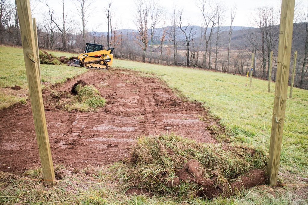 A small yellow skid steer sits on a patch of bare, freshly dug earth, surrounded by grassy field with wooden posts. Trees and hills are visible in the background under a cloudy sky.