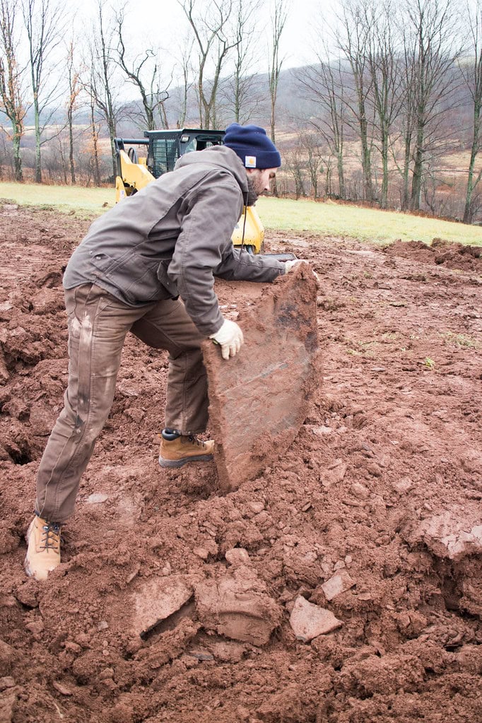 A person wearing a jacket, gloves, and boots lifts a large slab of dirt or clay in a muddy outdoor area, with bare trees and construction equipment in the background.