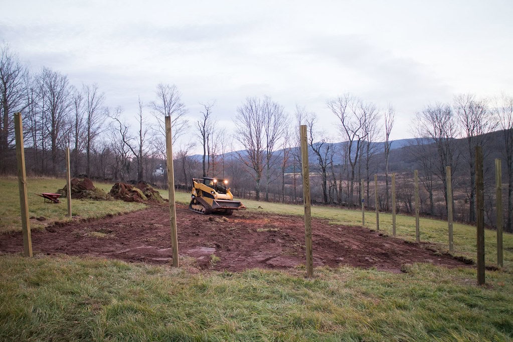 A small bulldozer levels dirt inside a rectangular area bordered by tall wooden posts on a grassy field with bare trees and hills in the background.