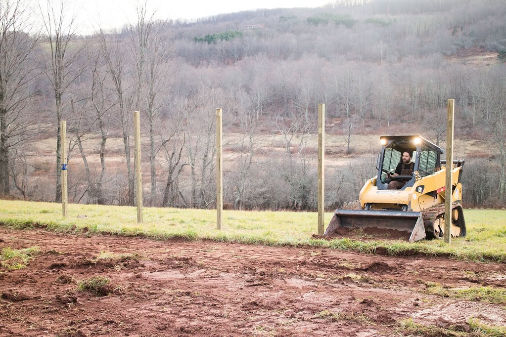 A person operates a small yellow bulldozer on grassy ground, driving between wooden fence posts on a rural property with hills and leafless trees in the background.