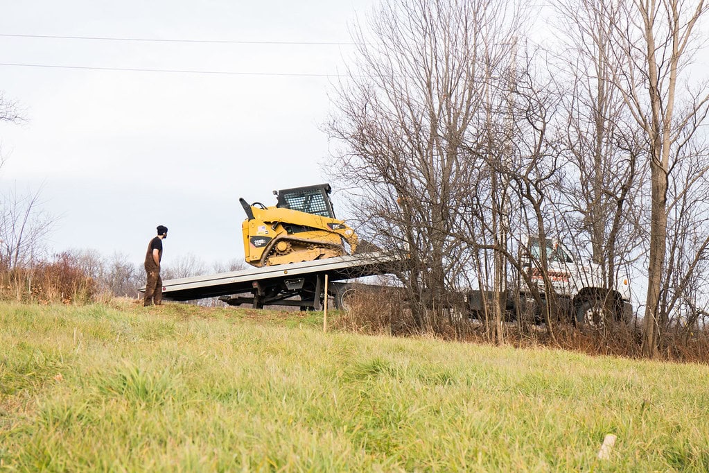 A person stands on grass, watching a yellow skid steer loader being loaded onto a flatbed truck ramp among leafless trees on a cloudy day.