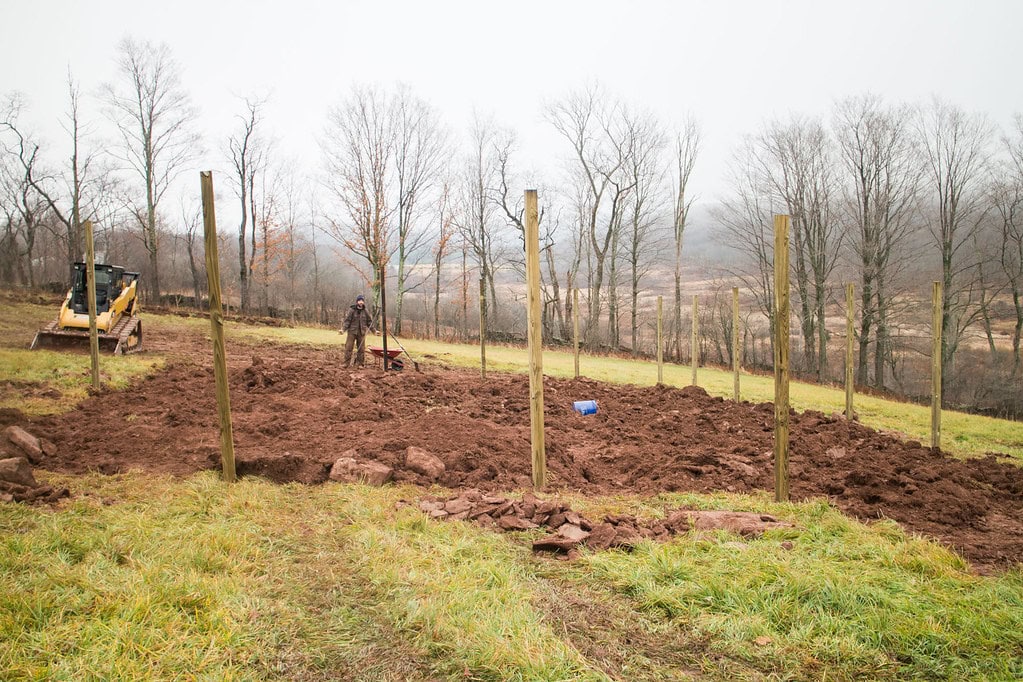 A person stands in a muddy field with wooden posts set in the ground. A wheelbarrow and construction equipment are nearby. Leafless trees and a foggy landscape are in the background.