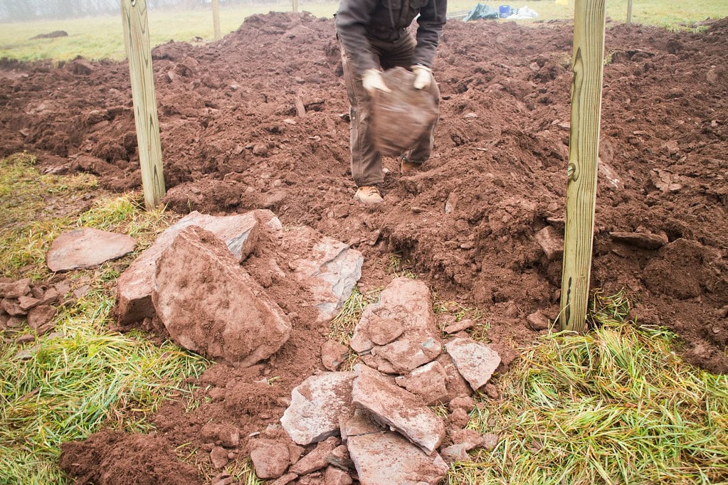A person is lifting a large rock in a muddy, freshly dug area surrounded by wooden posts. Broken rocks and patches of grass are visible in the foreground.