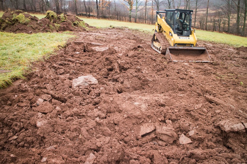 A yellow bulldozer is parked on a muddy, uneven construction site with churned earth and grassy edges, surrounded by trees in the background.