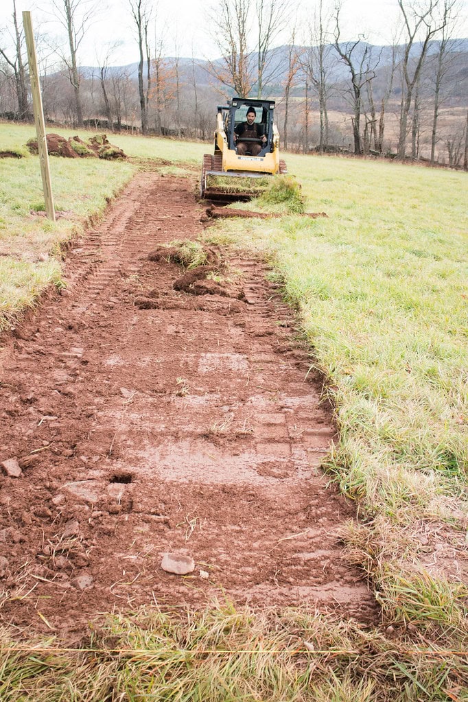 A person operates a small bulldozer, clearing a dirt path through a grassy field with bare trees and hills in the background.