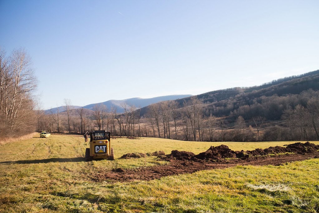 A construction vehicle digs a trench in a grassy field with trees and distant mountains under a clear blue sky.