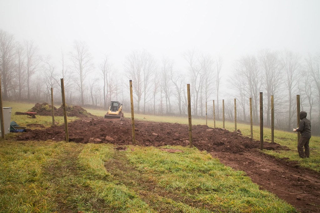 A person stands near a muddy field with upright wooden posts, while a small bulldozer works in the background on a foggy day. Bare trees line the horizon.