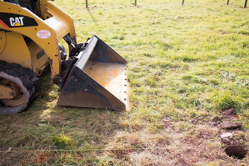 A yellow CAT skid steer loader with its bucket lowered sits on grassy ground next to a small dug hole.