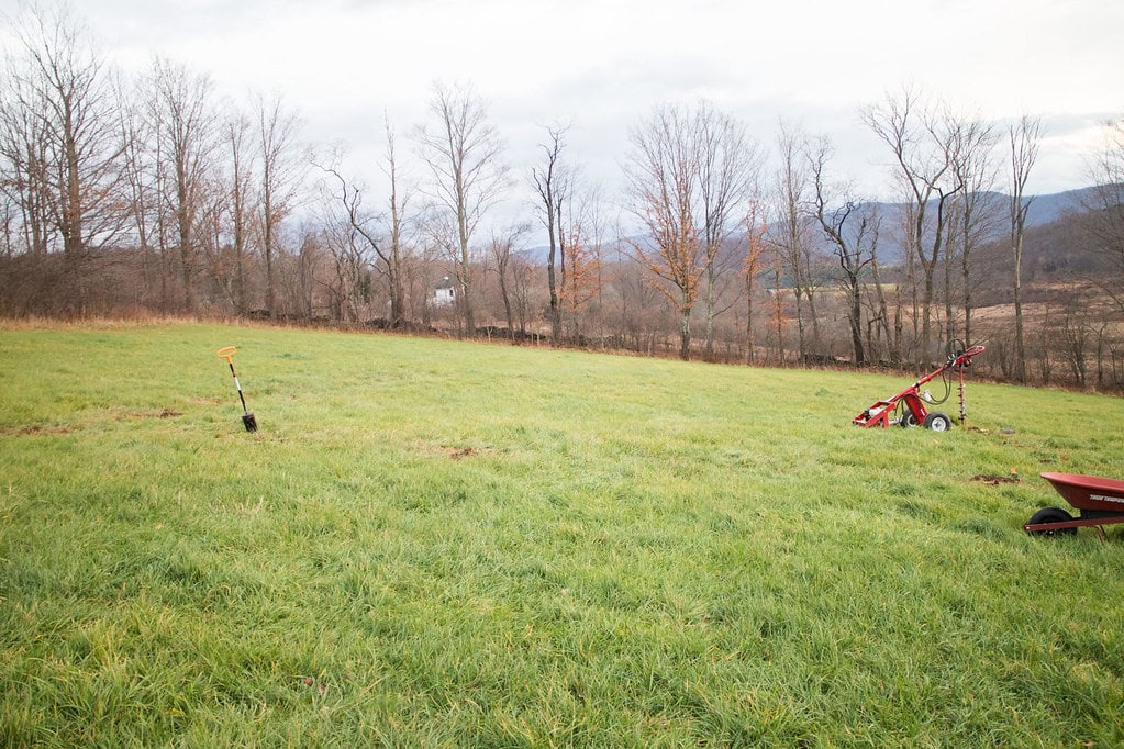 A grassy field with a shovel stuck in the ground, a red plow, and a wheelbarrow. Bare trees and distant hills are visible in the background under a cloudy sky.
