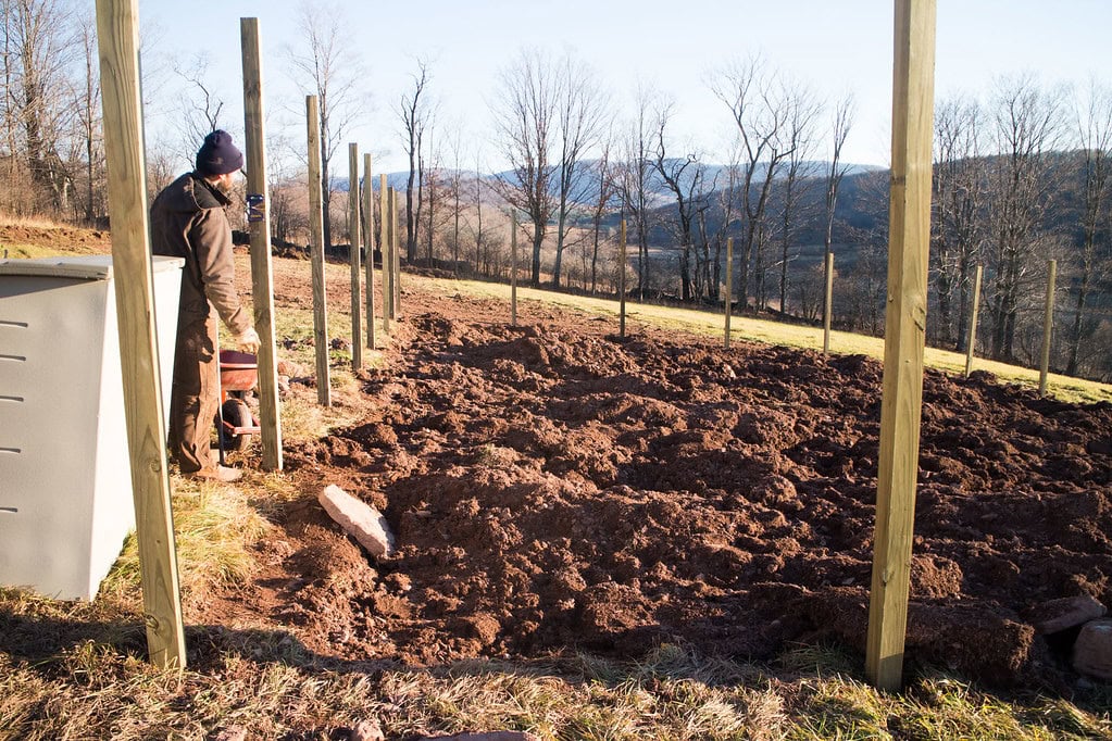 A person in work clothes stands next to wooden fence posts in a muddy field, with bare trees and hills visible in the background under a clear sky.