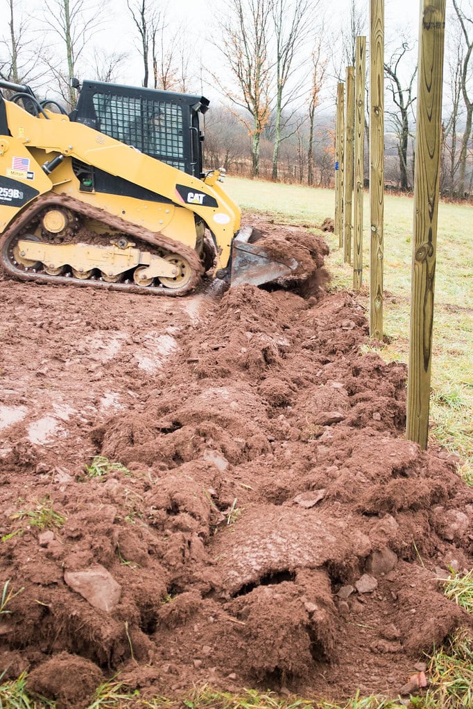 A yellow skid steer loader pushes dirt next to a row of wooden fence posts in a grassy field on a cloudy day. Bare trees are visible in the background.