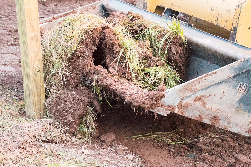 A close-up of a bulldozer blade lifting a chunk of grass and soil, revealing a large tunnel-like hole underneath the sod.