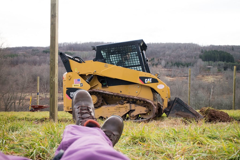 Person lying on grass, legs visible in foreground, facing a yellow CAT skid steer loader with tracks. The loader is parked on a grassy field with wooden posts and trees in the background.