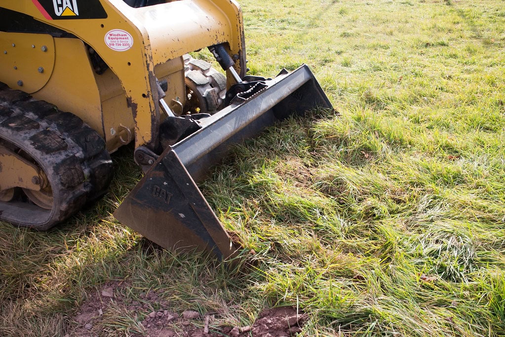 A yellow bulldozer with a front bucket is lifting up grass and dirt on a grassy field. Only part of the machine is visible on the left side of the image.