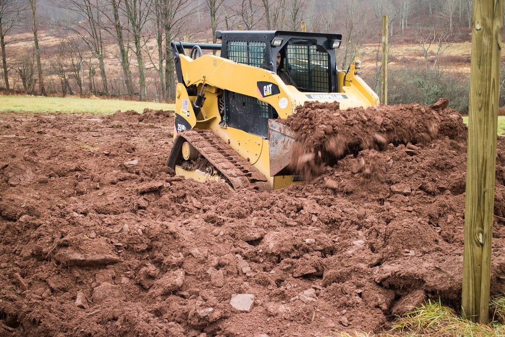 A yellow Caterpillar skid steer loader moves a pile of reddish-brown dirt on a muddy construction site, surrounded by bare trees and fields.