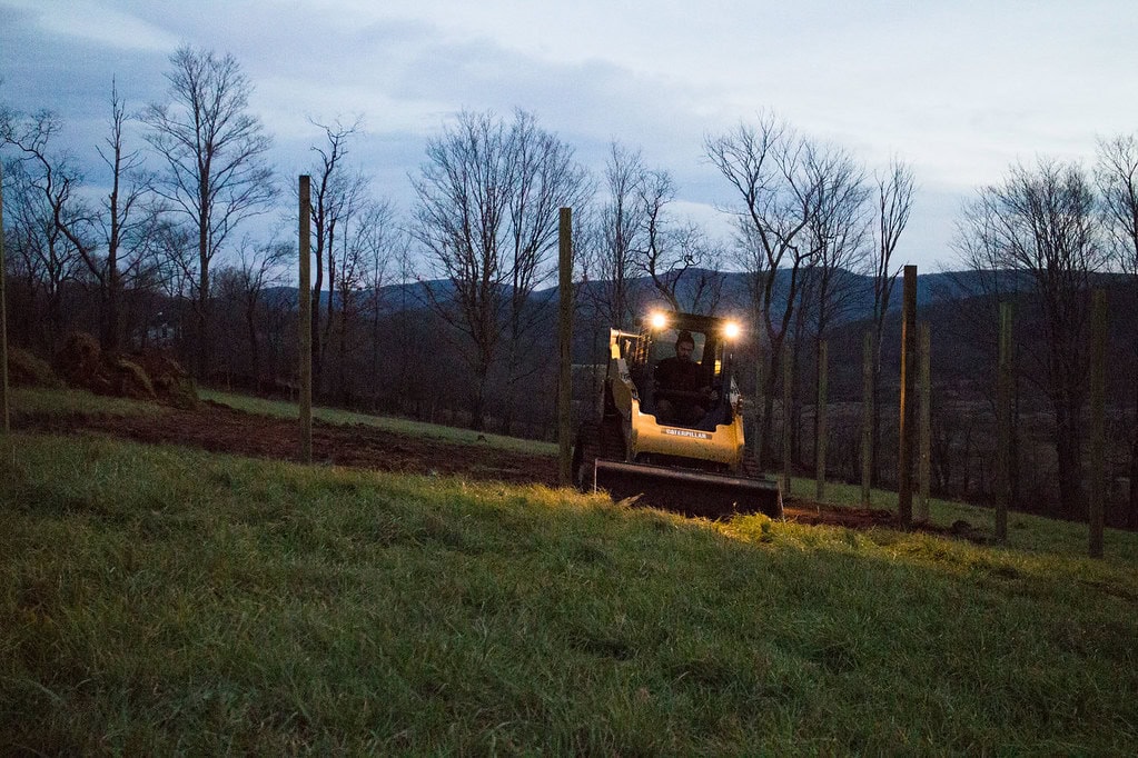 A small bulldozer with headlights on moves earth between tall wooden posts in a grassy field at dusk, with leafless trees and hills in the background.