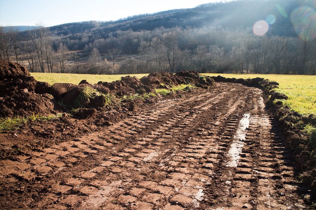 A dirt road with deep tire tracks runs through a grassy field, bordered by piles of soil, with leafless trees and hills in the background under sunlight.