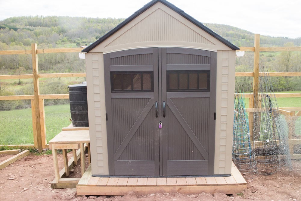 A small beige and brown garden shed with double doors, placed on a wooden platform in a fenced outdoor area, with fields and trees in the background.