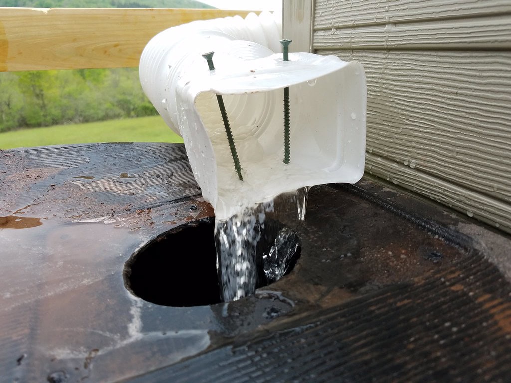 Water flows from a white plastic downspout, with two screws holding it in place, into a large black barrel through a circular hole. The setup appears to be part of a rainwater collection system.