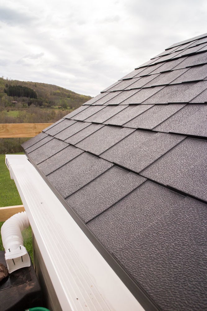 Close-up view of a shingled house roof with a white gutter system. In the background, grassy hills and trees are visible under a cloudy sky.
