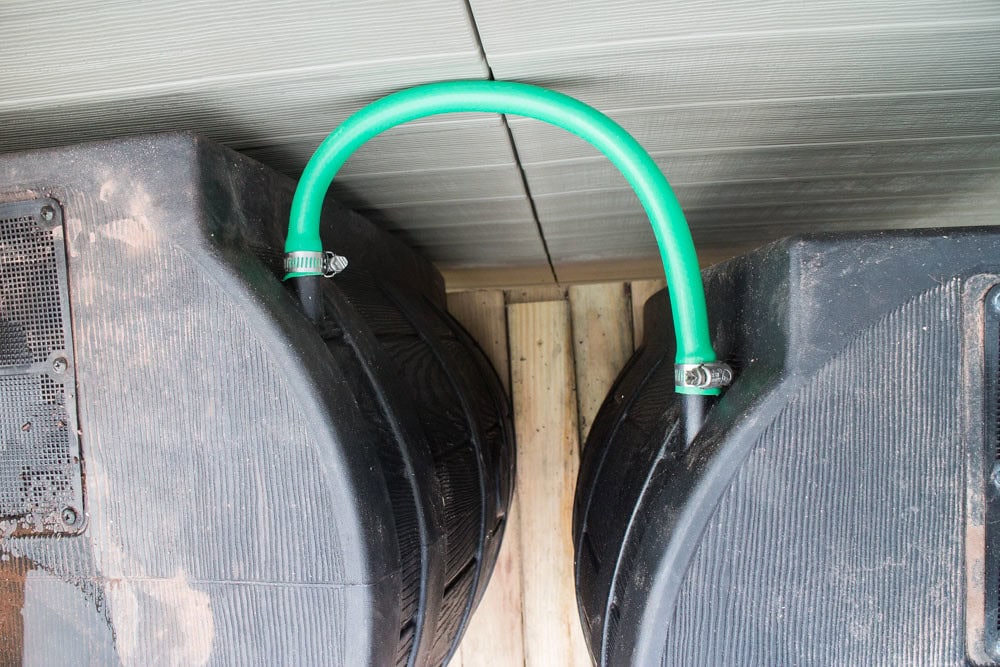 Two large black water tanks are connected at the top by a short green hose with metal clamps. The tanks are placed on a wooden platform under a corrugated roof.