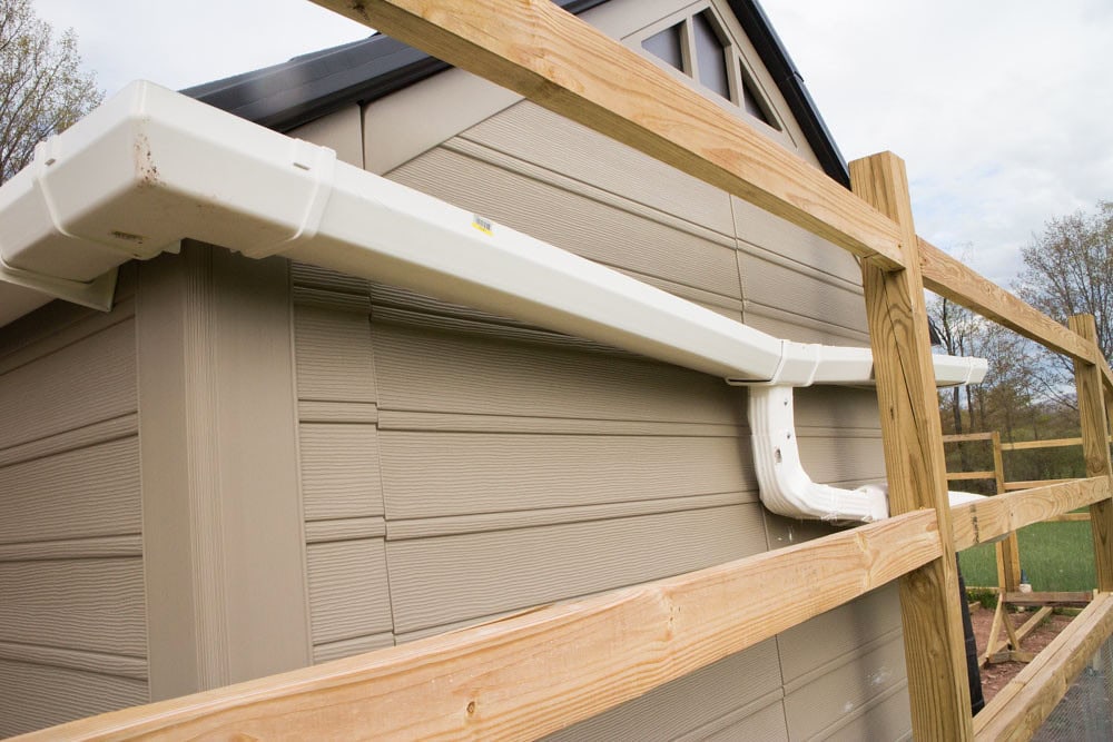White plastic rain gutter attached to the roof edge of a beige building, with wooden fencing running parallel to the structure. Trees and sky are visible in the background.