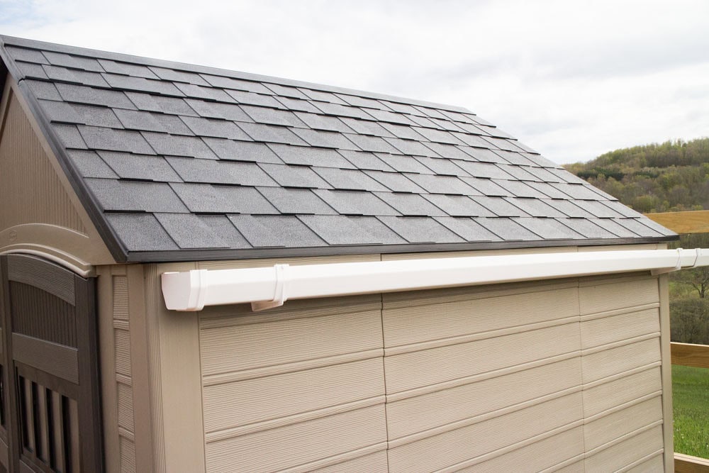 A beige outdoor shed with a black shingled roof and a white gutter attached, set against a grassy landscape with hills in the background.