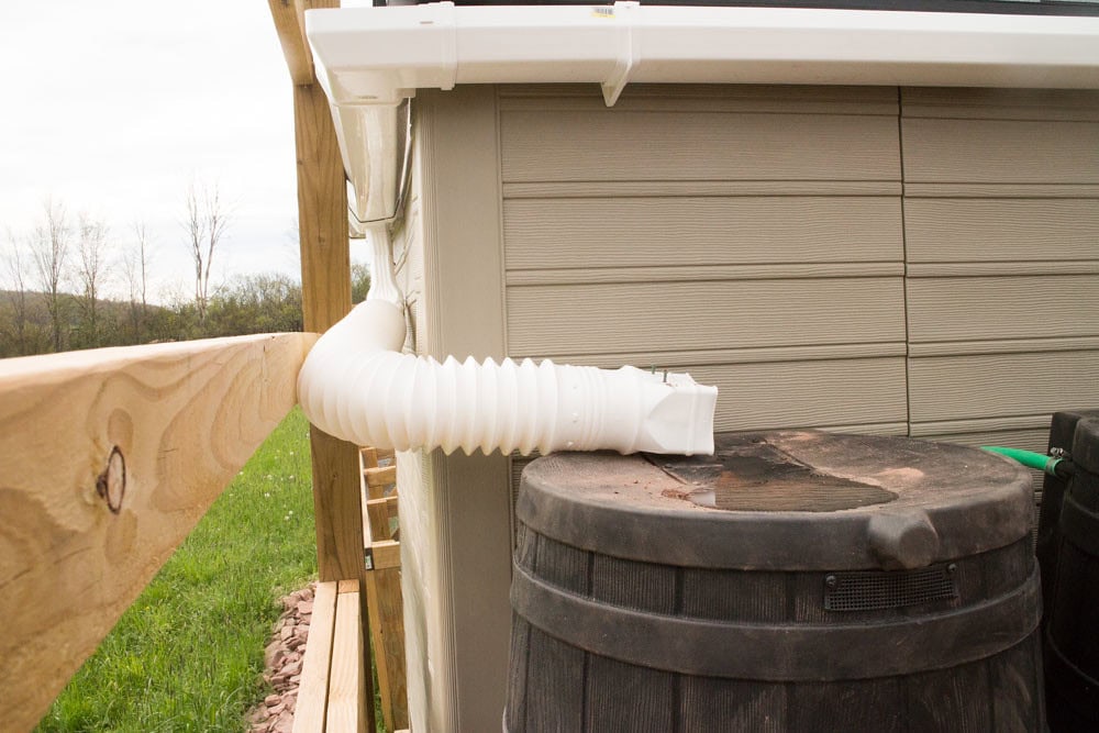 A flexible white downspout extension directs water from a gutter into a black rain barrel next to a beige building, with a wooden fence and grassy field nearby.