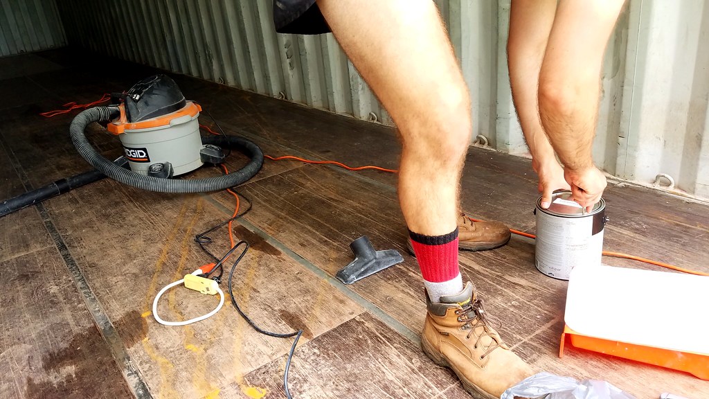 A person in tan boots and red socks opens a paint can on a wooden floor beside a vacuum, power cords, and cleaning supplies inside a metal container.